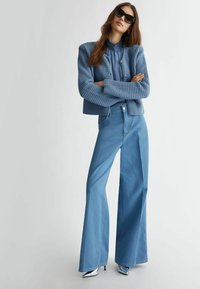 Giacca corta texturizzata blu chiaro sopra una blusa coordinata, abbinata a jeans a vita alta blu chiaro a palazzo e scarpe con tacco argentate lucide.