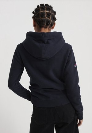 Osoba z krótką fryzurą bantu knot, ubrana w granatową bluzę z kapturem i czarne spodnie, stojąca z jedną ręką na biodrze, zwrócona tyłem.