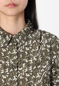 Grønt blomstermønster skjorte med krage, med hvite blomster og abstrakte blad design. Stoffet har en glatt tekstur.