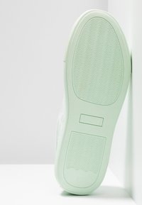 Białe sneakersy z jasnoniebieską gumową podeszwą, teksturowaną powierzchnią i wzorem fal. Obcas ma prostokątny akcent, ustawiony pod kątem do ściany.