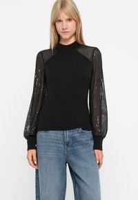Top negru cu croială ajustată și mâneci umflate, transparente, cu mărgele. Gulerul este înalt și se potrivește cu blugi largi de un albastru deschis.