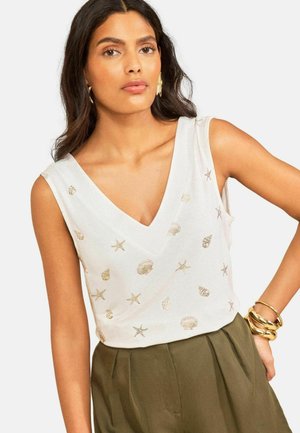 Femme portant un haut blanc sans manches avec des imprimés dorés de coquillages et d'étoiles de mer, pantalon taille haute olive, boucles d'oreilles dorées et plusieurs bracelets dorés.