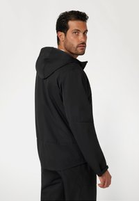 Juodas gobtuvu švarkas su matinio paviršiaus apdaila, turintis laisvą kirpimą, reguliuojamus rankogalius ir centrinę siūlę nugaros pusėje.