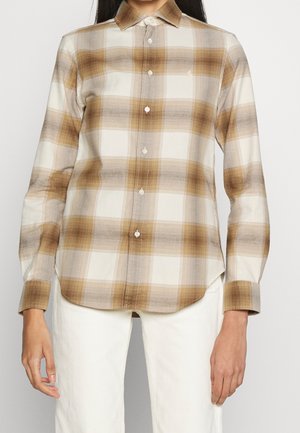 Femme portant une chemise à carreaux marron et beige à boutons avec manches longues et un pantalon blanc, debout devant un fond uni.