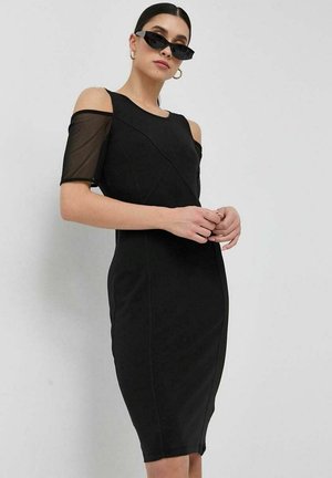 Abito nero con maniche corte trasparenti, silhouette aderente e dettagli con cuciture diagonali. La modella indossa occhiali da sole e si trova in piedi con le mani incrociate.