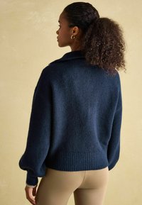 Marineblauer gestrickter Pullover mit geripptem Saum und übergroßen Ärmeln, mit einem Umlegekragen und strukturiertem Finish. Dazu sind bequeme beige Hosen sichtbar.