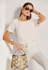 Beigefarbene Kurzarmbluse, weiße Jeans, hält eine Handtasche im Schlangenleder-Muster. Accessoires beinhalten große Sonnenbrillen und ein goldenes Armband.