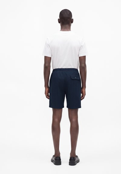 Homme portant un t-shirt blanc uni à manches courtes, un short bleu marine avec une poche arrière, et des chaussures noires, debout de dos sur fond blanc.