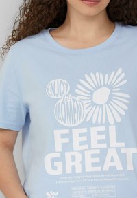 Lichtblauw katoenen t-shirt met witte tekst en een zonnebloemafbeelding. De tekst bevat "GENIET VAN HET MOMENT" en "VOEL JE GOED." Korte mouwen.