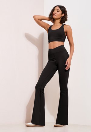 Kvinde med kort krøllet hår iført sort crop top og flagrende bukser, stående med en hånd bag nakken mod en lys væg.