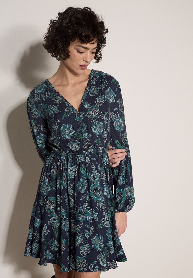 Femme aux cheveux bouclés portant une robe longue à manches longues à motif floral foncé, regardant vers le bas, debout contre un mur clair uni.