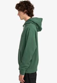 Felpa verde con cappuccio, tasca frontale, chiusura con cordoncino e polsini a costine. Tessuto morbido e casual con cuciture visibili.