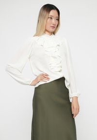 White blouse with ruffled front, long sleeves, and a high neckline, paired with a solid olive green satin skirt. Simple, elegant design.