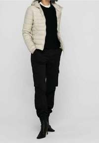 Doudoune beige clair avec capuche, pull noir en maille, et pantalon cargo noir avec poches latérales ; des bottines à talons noires complètent le look.