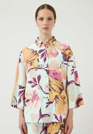 Kvinde iført løs, blomstret skjorte med knapper og matchende bukser i pastelnuancer, stående mod en ensfarvet lys baggrund.