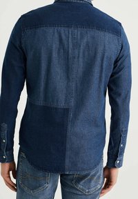 Mörk denimskjorta med en tvåfärgad design som har en enfärgad övre panel och en texturerad nedre del. Knappmanschetter och en rundad fåll som detaljer.