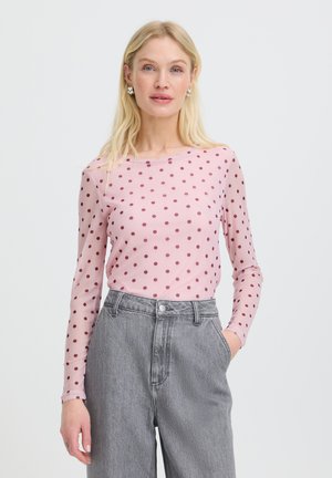 Femeie blondă purtând un top roz cu mâneci lungi cu buline închise la culoare și blugi gri cu talie înaltă, stând cu o mână în buzunar.