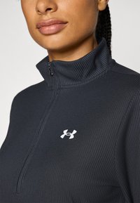 Crna majica s puloverom na zip s rebrastom teksturom. Ima mali bijeli logo na lijevoj strani prsa i visoki ovratnik.