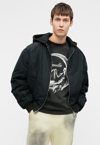 Fekete bomberdzseki kapucnival, cipzárral, bordás mandzsettákkal. Szürke grafikus póló kör alakú mintával. Világos bézs nadrág ellipszis textúrával.