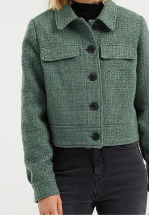 Grøn tekstureret jakke med knapper og klaplommer på brystet, båret over en sort top og kombineret med sorte jeans med høj talje.