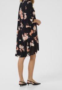 Femme portant une robe noire à fleurs de longueur genou avec des manches 3/4 et des chaussures à talons bas à bride arrière noires, se tenant devant un fond blanc.