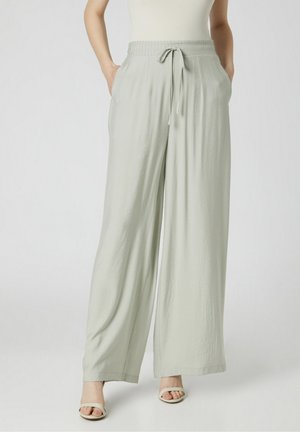 Kobieta w jasnoszarych szerokich spodniach z elastycznym pasem i sznurkiem, w białych sandałach z odkrytymi palcami, z rękami w kieszeniach.