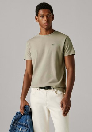 Õliveroheline puuvillane T-särk lühikeste varrukatega, ümar kaelus ja väike must logo rinnal, kombineeritud valgete pükste ja sinise teksatööriietusega.