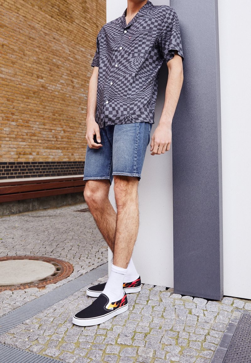 Personne appuyée contre un mur portant une chemise à manches courtes à carreaux noirs et gris, un short en denim, des chaussettes blanches et des chaussures noires à enfiler avec un motif de flammes.