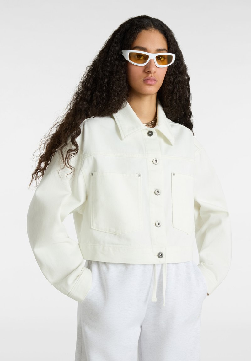 Witte cropped denimjas met knopen aan de voorkant, twee borstzakken en een klassieke kraag, gecombineerd met grijze broek en witte zonnebril.