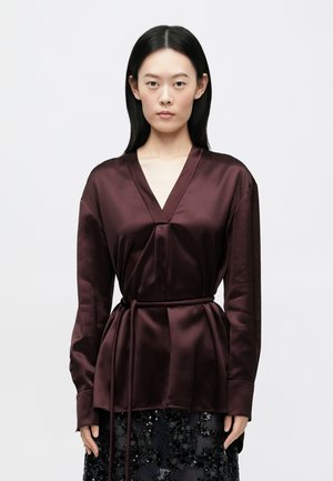 Viininpunainen satiinipusero, jossa on V-aukko, pitkät hihat ja vyötärön solmittava nauha. Sileä pinta ja yksinkertainen muotoilu ilman kuvioita.