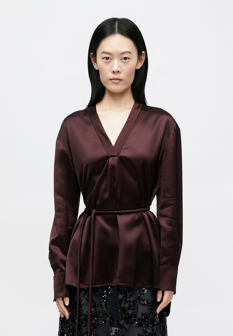 Μπλούζα από σατέν σε μπορντό, με V-λαιμόκοψη, μακρ�ιά μανίκια και ζώνη στη μέση. Ομαλή υφή και απλός σχεδιασμός χωρίς σχέδια.