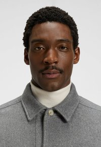 Gray herringbone wool coat with a button closure and a pointed collar, worn over a cream turtleneck. Smooth texture and neat finish.