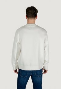 Weißer Sweatshirt mit rundem Hals, langen Ärmeln und gerippten Bündchen. Kombiniert mit blauen Jeans, von hinten betrachtet. Glatte Stoffoberfläche.