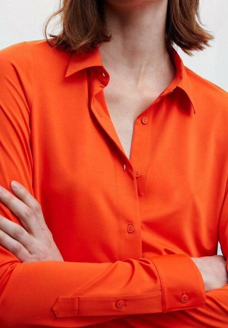 Blouse ajustée à manches longues de couleur orange vif, confectionnée dans un tissu lisse. Dotée d'un col classique et d'une fermeture boutonnée à l'avant avec des boutons assortis.