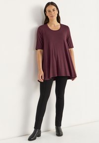 Kvinna står mot en vägg klädd i en lös burgundy tunika, svarta smala byxor och svarta ankelboots.