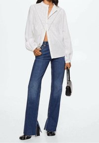 Weißes, langärmliges Hemd mit Knopfleiste aus strukturiertem Stoff und Stickerei. Kombiniert mit hoch taillierten, ausgestellten blauen Jeans mit Schlitzsäumen.