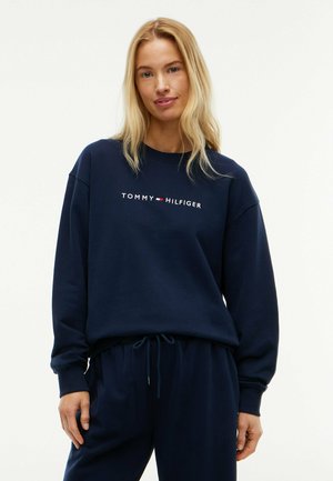 Lyshåret kvinde iført en navyblå Tommy Hilfiger sweatshirt og matchende navyblå bukser, der står mod en hvid baggrund.