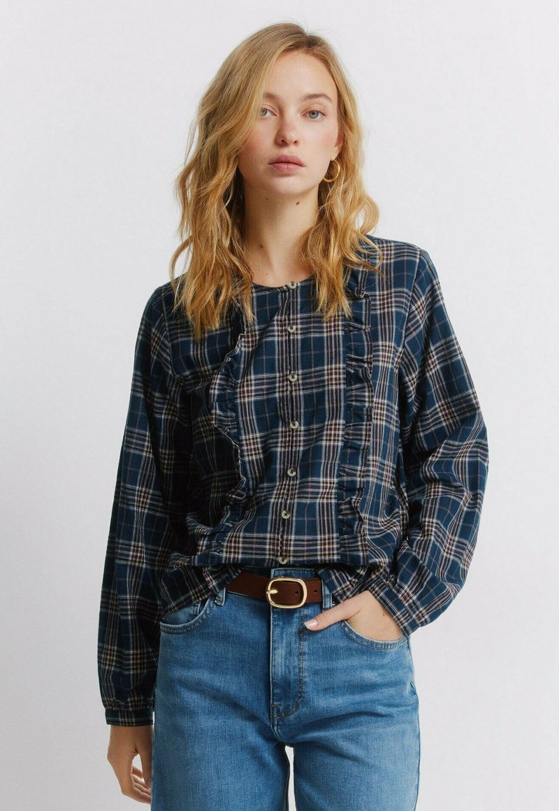 Marineblauw geruit overhemd met ruches en knoopsluiting. Lange mouwen en een losse pasvorm. Gecombineerd met een hooggesneden blauwe jeans en een bruine riem.