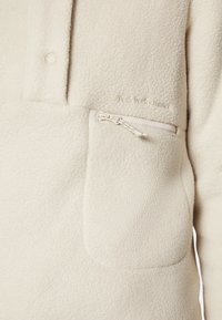 Fleece pullover i lys beige med halv-snaps krave, lynlåslomme og præget brandlogo. Blød tekstur og minimalistisk design.