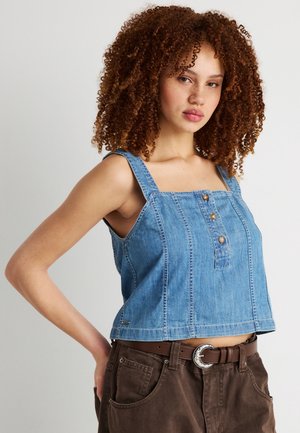 Junge Frau mit lockigem Haar, die ein ärmelloses blaues Jeansoberteil und braune Hosen mit Gürtel trägt, steht vor einem einfarbigen Hintergrund.