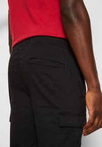 Černé pracovní kalhoty s elastickým pasem, postranními kapsami a plochými zadními kapsami. Látka se jeví jako měkká s hladkou texturou.