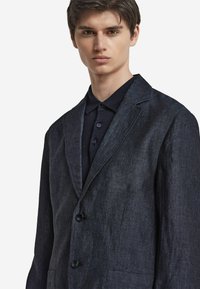 Mörkblå linnet blazer med texturerad finish, enkelknäppt design, spetsiga slag och två framfickor, bärs över en mörk polo skjorta.