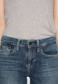 Midja klädd i blekta blå denimjeans med knäppning och dragkedja stängd, tillsammans med en instoppa ljusgrå ribbad topp.