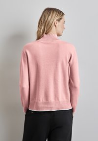 Rosaner, gestrickter Pullover mit hohem Rippenkragen, langen Ärmeln und geripptem Saum, getragen über schwarzen Hosen und zeigt eine weiche Textur.