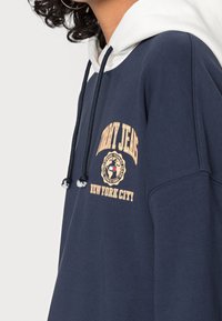 Námořnická mikina s kapucí a bílým podšitím, s vyšitým logem "TOMMY JEANS" a designem kulatého odznaku na hrudi. Akcenty na stahovací šňůrce.