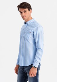 Camisa a rayas azul claro con cuello button-down, bolsillo frontal y logo. Hecha de algodón, cuenta con mangas largas y un dobladillo redondeado.