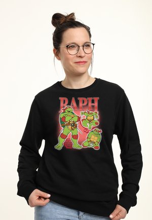 Femme portant des lunettes et un sweat-shirt noir avec trois personnages des Tortues Ninja et le texte "RAPH" en rouge.