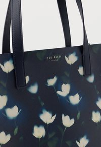 Sac fourre-tout bleu marine à motif floral avec une finition texturée, présentant des motifs de fleurs blanches et un logo doré "Ted Baker London", complété par des anses robustes.