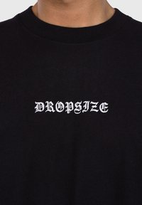 Svart bomullssweatshirt med rund hals og hvit brodert tekst "DROPSIZE" i en dekorativ skrifttype.