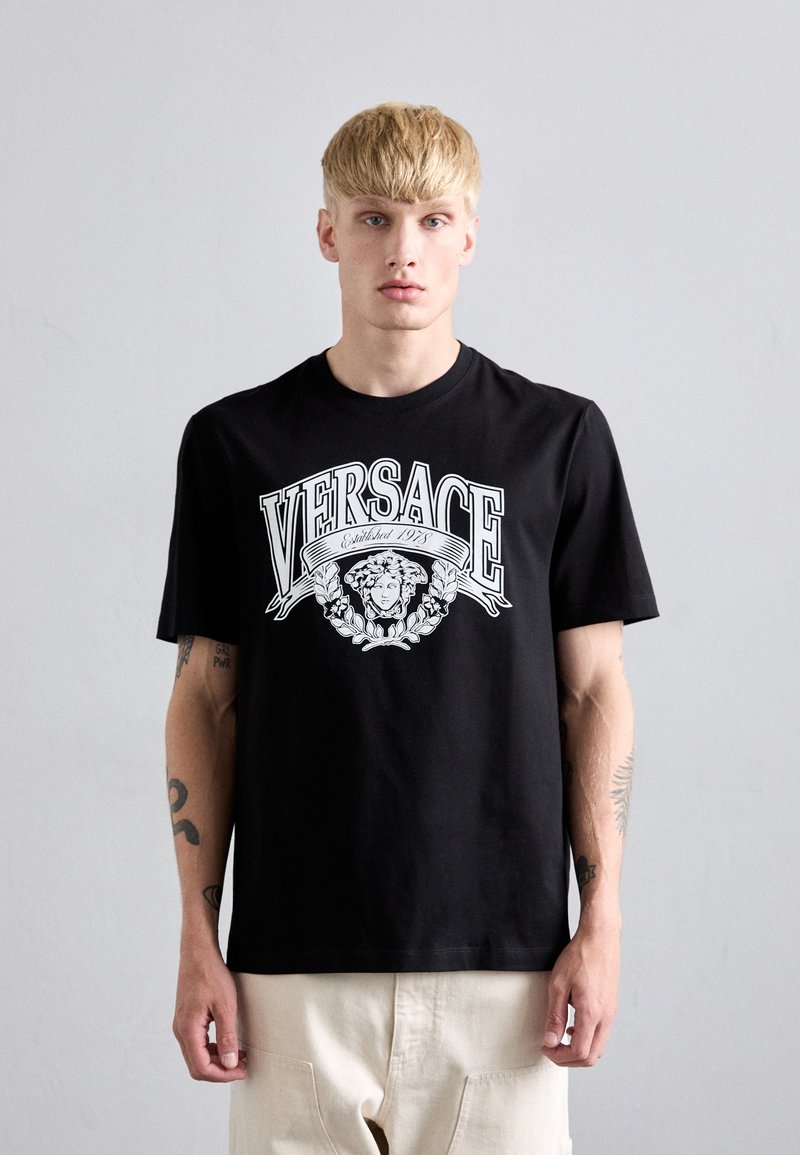 Juodas medvilnės marškinėliai su baltu grafiniu logotipu, kuriame pavaizduota liūtas, užrašas "VERSACE" ir gėlių akcentai. Trumpomis rankovėmis, laisvas kirpimas.